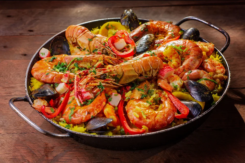 BookNoFees: Esimerkkikuva: Pannu mereneläviä sisältävää paellaa, jonka päällä on äyriäisiä ja punaisia paprikoita