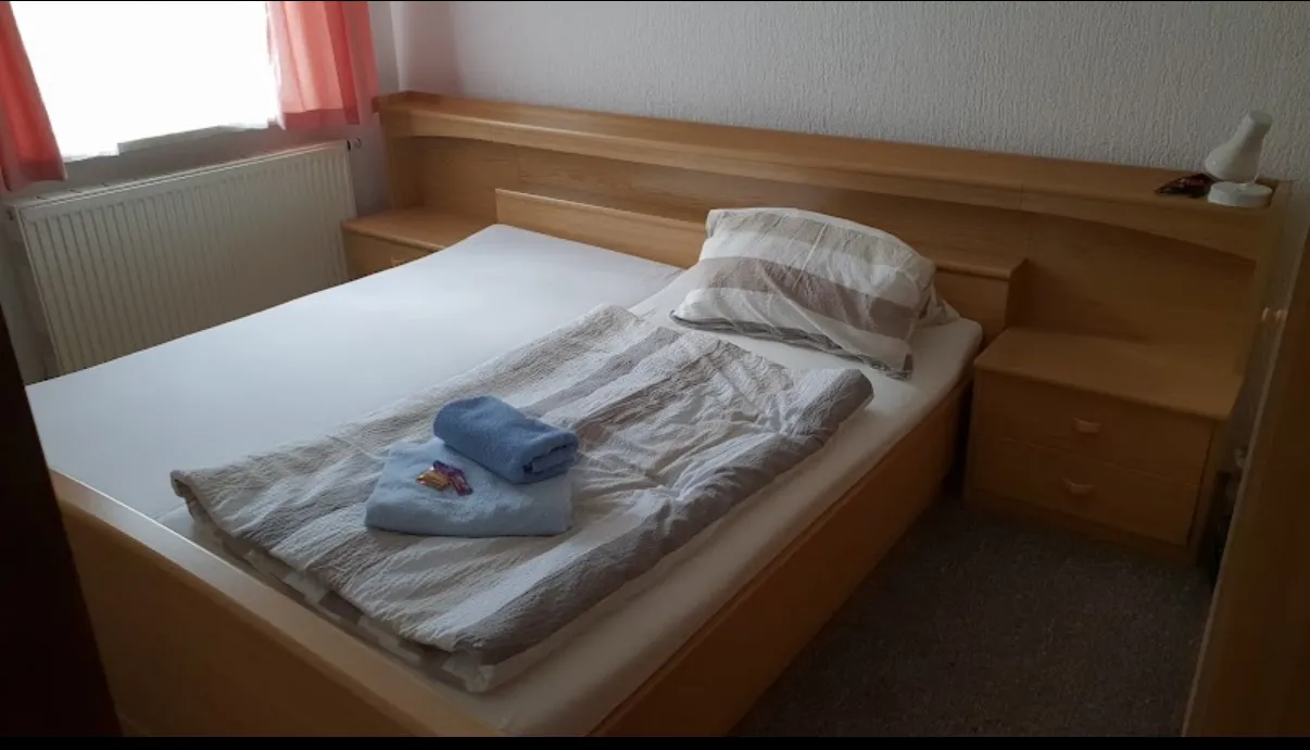 BookNoFees: semesterboende i Schönfeld, Sachsen, Tyskland – Sovrum med sängram i trä, vikta handdukar och ett fönster med rosa gardiner