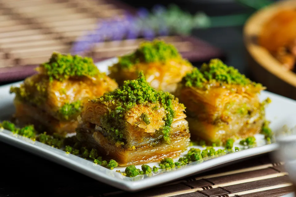 BookNoFees: Esimerkkikuva: Neljä kerrosta baklava-leivonnaista, joiden päällä on murskattuja pistaasipähkinöitä, valkoisella lautasella